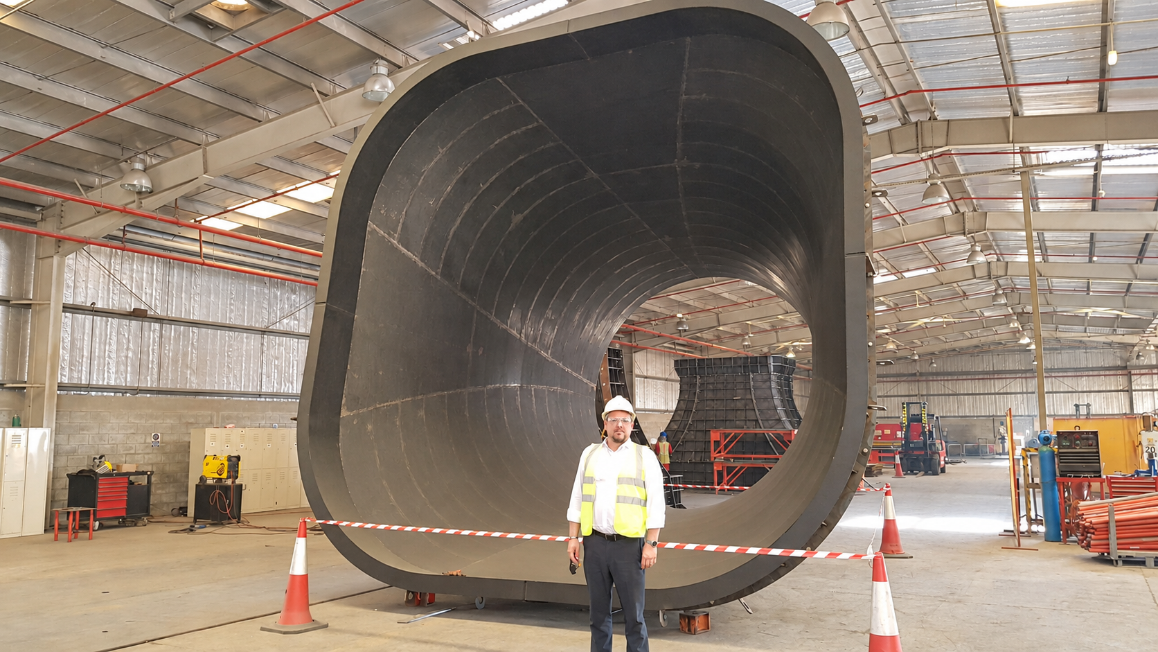 Centin Baxter wearing a hard hat and high-visibility vest beside a large curved steel form inside the special steel forms factory